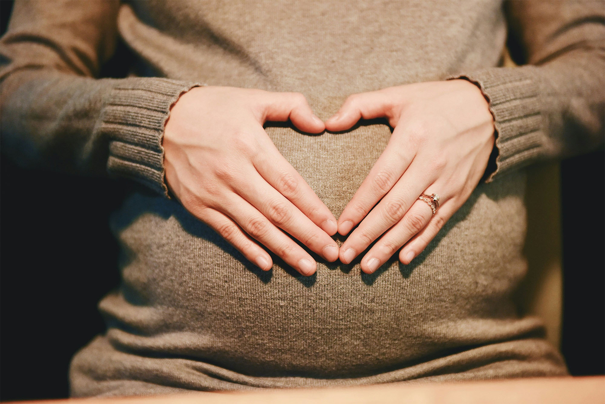 Expectant mother in sweater showcasing a heart hand gesture over her belly, highlighting the beauty of pregnancy.